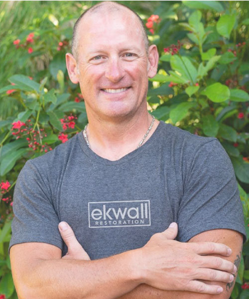Smiling man wearing an Ekwall Restoration t-shirt, standing in front of green foliage, representing the company's commitment to quality home restoration services.