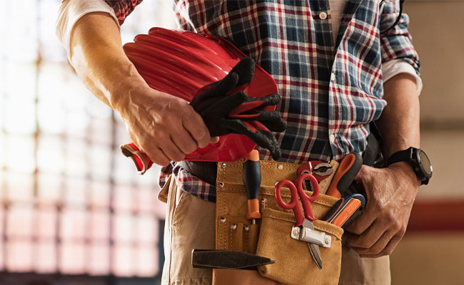 Person wearing a plaid shirt and tool belt, holding a hard hat and gloves, ready for home restoration and repair work in Payson.